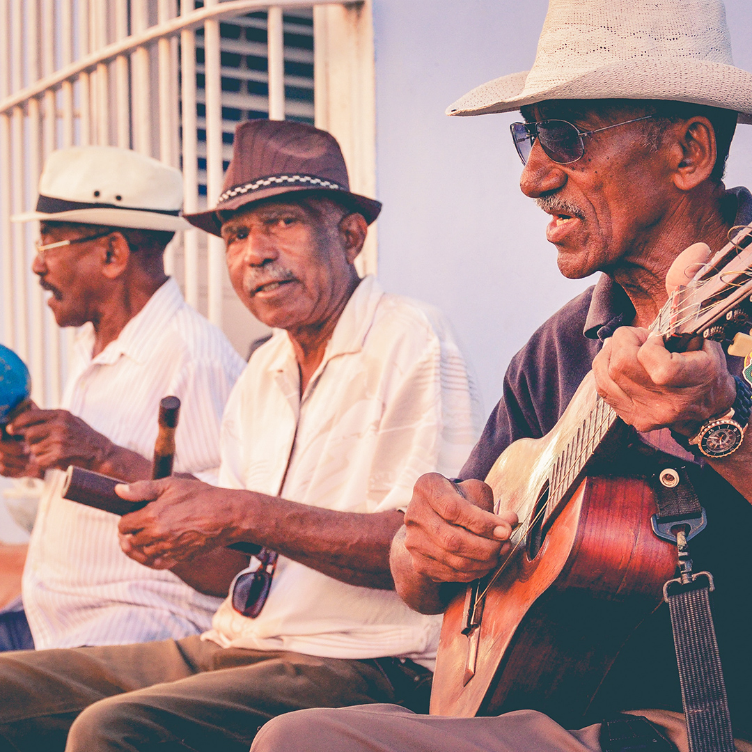 Tres adultos mayores tocando musica usando instrumentos tipicos puertorriqueños.